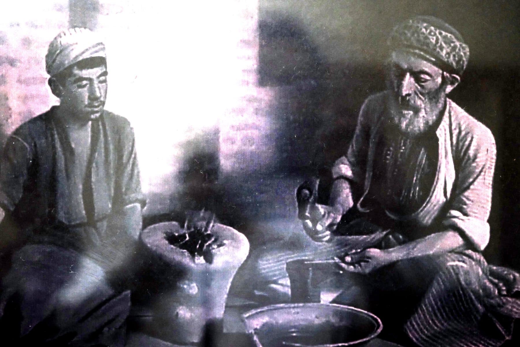 Photograph of an elderly goldsmith and a young assistant beside a clay forge