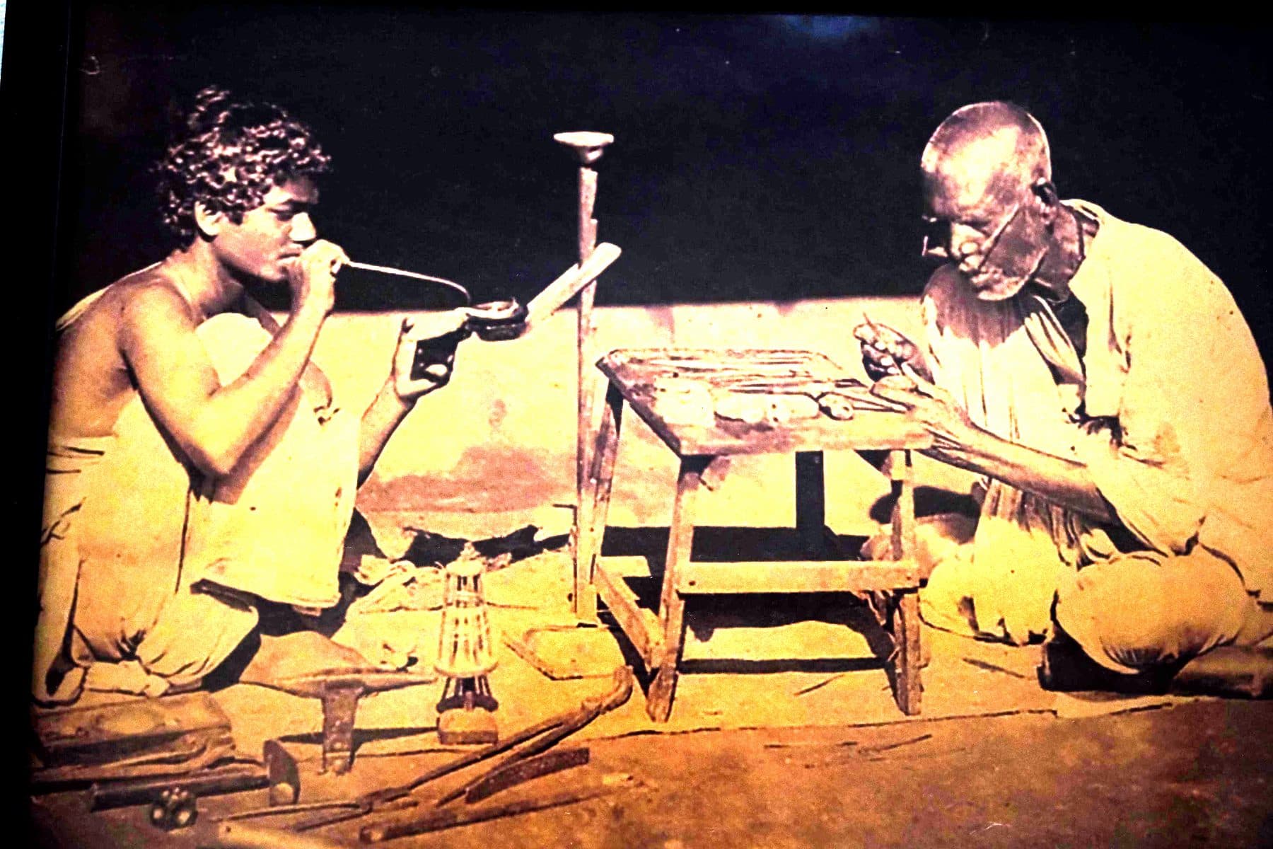 Sepia photograph of two goldsmiths at a low bench with an oil lamp and stake anvil